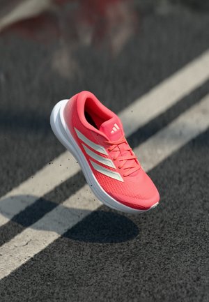 Chaussure de sport corail vif avec des rayures blanches en plein air au-dessus d'une route asphaltée avec une ligne blanche, projetant une ombre en dessous.