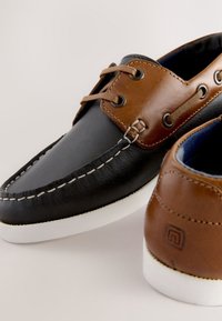 Chaussures bateau en cuir bicolores avec lacets marron, coutures blanches contrastantes et semelles en caoutchouc blanches sur fond clair.