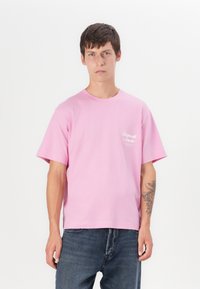 Pink bomulds T-shirt med rund halsudskæring og korte ærmer, med et hvidt logo trykt på øverste venstre side. Båret med blå jeans.