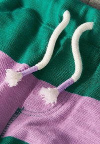 Zelený a fialový pruhovaný materiál s bílými stahovacími šňůrkami, vyrobený z texturovaného pleteného materiálu a zakončený třásněmi. Představuje ležérní, sportovní design.