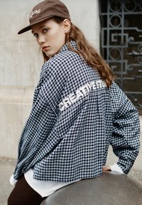 Camicia a quadri blu navy e bianca oversize con "CREATIVESTUDIOS" ricamato sul retro, abbinata a un cappellino marrone. Texture liscia.