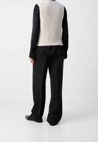 Eine Person trägt ein schwarzes Langarmshirt, schwarze weite Hosen, schwarze spitze Schuhe und eine ärmellose beige strukturierte Weste und steht mit dem Rücken zur Kamera.