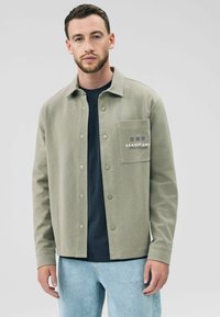 Khaki button-up jacket made of textured fabric, featuring a chest pocket with embroidered text and a collared neckline, paired with a dark shirt.