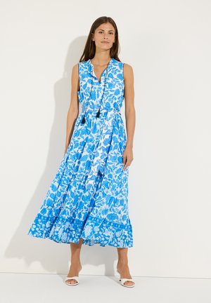 Femme portant une robe midi fleurie bleue et blanche sans manches avec un col en V et des liens à pompons, associée à des sandales blanches à talons.