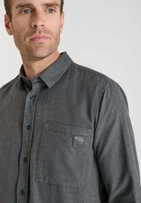 Camisa de franela gris con cuello botón, botones negros, un bolsillo en el pecho y un acabado texturizado. Presenta un parche de logo en el bolsillo.