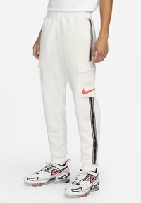 White jogger pants with side cargo pockets, black and red stripe detailing, and a red Nike logo on the thigh. Worn with multi-colored sneakers.