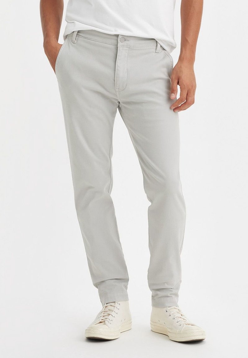 Homme portant un pantalon slim gris clair avec des poches avant et des baskets blanches montantes, se tenant avec une main dans la poche.