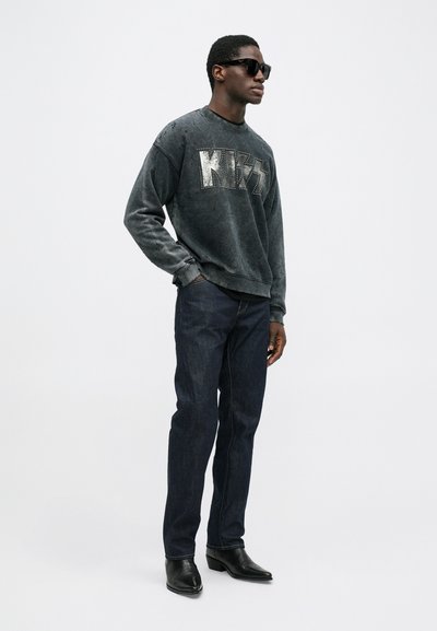 Tumehall sweatshirt, millel on suurelt kirjas "KISS" metallikaktsentidega, koos tumedate teksade ja mustade terava ninaga saapadega ning prillidega.