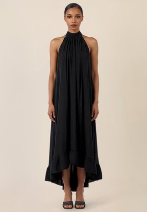 Frau steht und trägt ein ärmelloses schwarzes Midikleid mit Neckholder, Stufenrock und schwarzen offenen Absatzschuhen vor einem einfarbigen beigen Hintergrund.