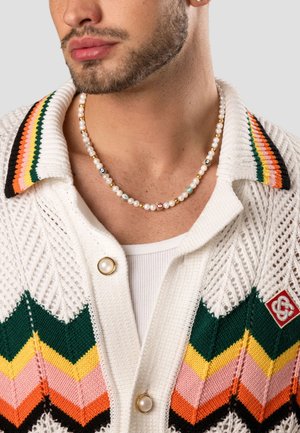 Halskæde med hvide perler, farverige accentperler og guldsmykker, båret sammen med en hvid strikket cardigan med multikolorede zigzag-mønstre.