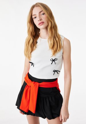 Joven con cabello largo y rubio que lleva un top sin mangas blanco con lazos negros, falda negra y un suéter rojo atado alrededor de la cintura.