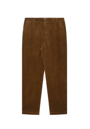 Junger Mann steht mit Händen in den Taschen, trägt ein gestreiftes Poloshirt, hochgekrempelte braune Hose und dunkelbraune Loafers vor beigem Hintergrund.