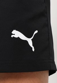 Logo Puma blanc sautant sur un short de sport noir porté sur la partie supérieure de la jambe d'une personne.