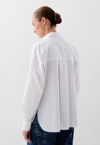 Camicia bianca a maniche lunghe con colletto classico, dotata di yoke posteriore, dettagli a pieghe e spacchi laterali, indossata con jeans blu scuro.