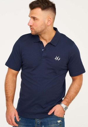 Marineblaues Poloshirt aus Baumwolle mit klassischem Kragen, kurzen Ärmeln und kleinem weißem Logo auf der Brust. Jeans aus Denim und eine silberne Uhr sind sichtbar.