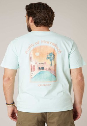 Homme portant un T-shirt bleu clair avec un motif estompé représentant une piscine, un palmier et le soleil, avec l'inscription « Oasis de Marrakech » au dos.