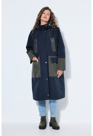 Kiharatukkainen nainen pukeutuneena pitkään, tummansiniseen ja oliivinvihreään vedenpitävään takkiin, vaaleansiniin farkkuihin ja oliivinvihreisiin nauhallisiin saappaisiin, seisoo valkoista taustaa vasten.