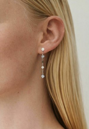 Prim-plan cu urechea unei femei blonde care poartă o cercei cu lanț argintiu și mărgele mici albastre și albe.