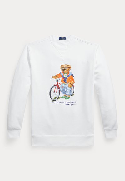 Fehér hosszú ujjú pulóver egy medvét ábrázoló grafikával, amely napszemüveget, narancssárga dzsekit, farmert és zöld tornacipőt visel, piros kerékpár mellett áll.