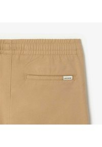 Beige šorti ar elastīgu jostu un aizmugurēju kabatu, kurā ir balts Lacoste zīmols. Audums izskatās gluds ar pielāgotu dizainu.
