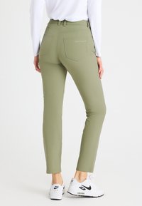 Olijfgroene fitted broek met een hoge taille en twee achterzakken, gecombineerd met witte sneakers met een zwart logo. Gladde textuur.
