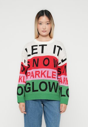 Jeune femme portant un pull multicolore avec les mots "LAISSEZ NEIGER", "ÉCLAT" et "LUMIÈRE", associé à un jean bleu, se tenant devant un fond blanc.