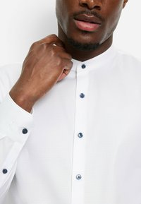 Weißes Hemd mit Stehkragen, ausgestattet mit blauen Knöpfen und einer dezenten Textur. Manschetten-Detail sichtbar mit passendem blauen Knopf.