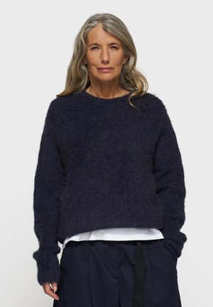 Mørkeblå fluffy sweater med rund hals, nedfaldne skuldre og brede manchetter, lagd over en hvid skjorte. Blød tekstur og afslappet pasform.