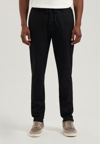 Schwarze Jogginghosen aus weichem Material, mit einem Kordelzugbund, seitlichen Taschen und schmal zulaufenden Beinen. Getragen mit grauen Turnschuhen.