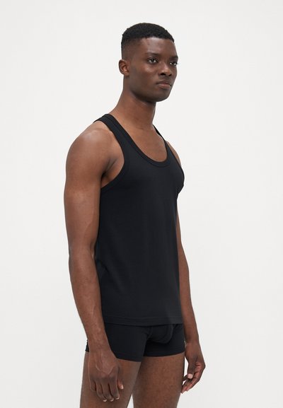 Junger Mann mit kurzen Haaren, der ein eng anliegendes schwarzes Tanktop und schwarze Boxershorts trägt und vor einem schlichten weißen Hintergrund steht.