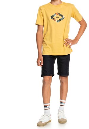 T-shirt jaune avec un logo floral en forme de diamant, associé à un short en denim sombre. Baskets blanches avec des accents gris et des chaussettes rayées.