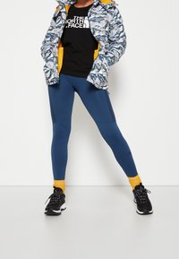 Modelka na sobě má modrou vzorovanou bundu se žlutým lemováním, černé tričko The North Face, modré legíny, žluté ponožky a černé běžecké boty.