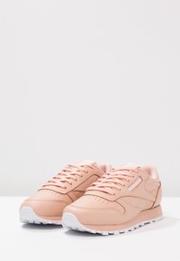 Ett par pastellrosa Reebok-sneakers med vita sulor, detaljerad sömnad och logotyp på sidan, placerade på en neutral vit yta.