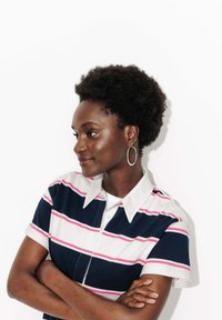 Polo-shirt avec col, rayures horizontales bleu marine, blanc et rose. Dispose de manches courtes et d'une fermeture à boutons à l'avant. Le modèle porte des boucles d'oreilles créoles.