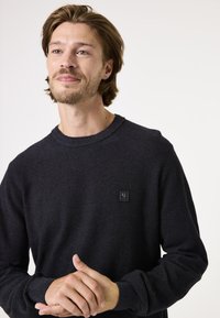Zwarte gebreide trui met een gestructureerd chevronpatroon, ronde hals en een klein merkembleem op de borst. Model poseert met handen gevouwen.