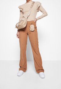 Beige gerippte Hose mit ausgestellter Form, kombiniert mit einem gestreiften Langarmoberteil und einer beigen Kreuzbody-Tasche. Weiße Sneaker vervollständigen das Outfit.