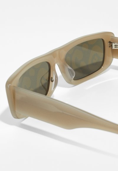 Des lunettes de soleil avec une monture en plastique beige, des verres teintés verts et un motif texturé sur la surface intérieure des branches pour un détail supplémentaire.