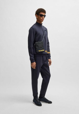 Black crossbody bag with gold accent, navy sweatshirt and joggers, textured fabric, sleek design, worn with sunglasses and dark sneakers.