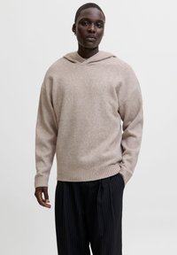 Suéter de punto beige con capucha, que presenta un corte relajado y puños acanalados. Combinado con pantalones negros de rayas para un contraste.