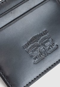 Κοντινό πλάνο πορτοφολιού από μαύρο δέρμα Levi Strauss & Co. με ανάγλυφο λογότυπο που δείχνει δύο άλογα να τραβούν ένα ζευγάρι τζιν.