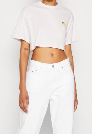 Osoba ubrana w biały krótki t-shirt z małym żółtym detalem cytryny i białe jeansy z wysokim stanem, pokazująca brzuch i tatuaże na ramionach.