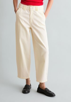 Osoba nosząca kremowe spodnie z wysokim stanem i szerokimi nogawkami, czarne mokasyny oraz włożoną do środka czerwoną bluzkę, stojąca z rękami w kieszeniach.