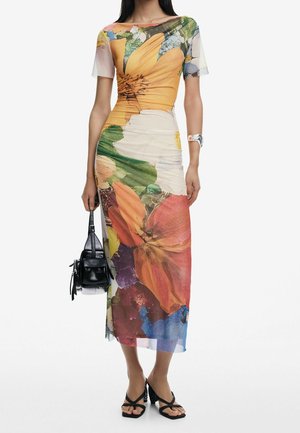 Frau trägt ein figurbetontes, buntes Midikleid mit Blumenmuster und kurzen Ärmeln, hält eine kleine schwarze Handtasche und trägt schwarze Sandalen mit Absatz.