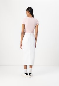 Lys rosa t-skjorte som sitter tett, kombinert med et langt hvitt denimskjørt. Modellen har på seg hvite sneakers med svarte detaljer og lysegrå sokker.