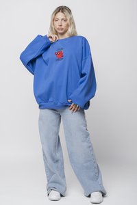 Zila, pārāk liela izmēra sporta krekls ar mazu sarkanu multfilmu dizainu uz krūtīm, saskaņots ar gaiši zilu plašu biksēm un baltām kedām.