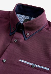 Camisa de botón color marrón con una textura tejida, forro azul en el cuello y detalle de bolsillo estampado. Cuenta con cinco botones y un bolsillo plano.