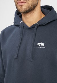 Felpa blu navy in misto cotone, con cappuccio regolabile e un logo bianco ricamato sul petto. Tessuto liscio.