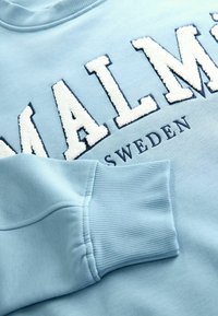 Ljusblå hoodie med "MALMÖ" i vit fluffig text med marinblå detaljer och ribbad mudd. Mjuk materialstruktur synlig.