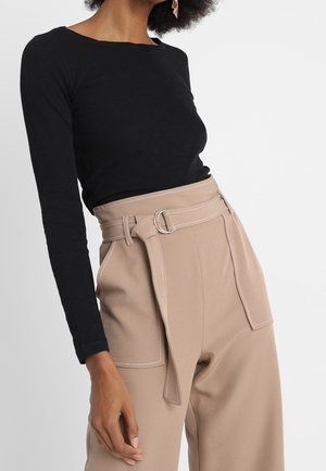 Vrouw draagt een zwarte top met lange mouwen in hoge beige broek met grote voorzakken en een bijpassende riem.