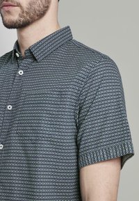 Uomo che indossa una camicia a maniche corte con un motivo geometrico scuro e colletto a punta, mostrato dalle spalle in su.
